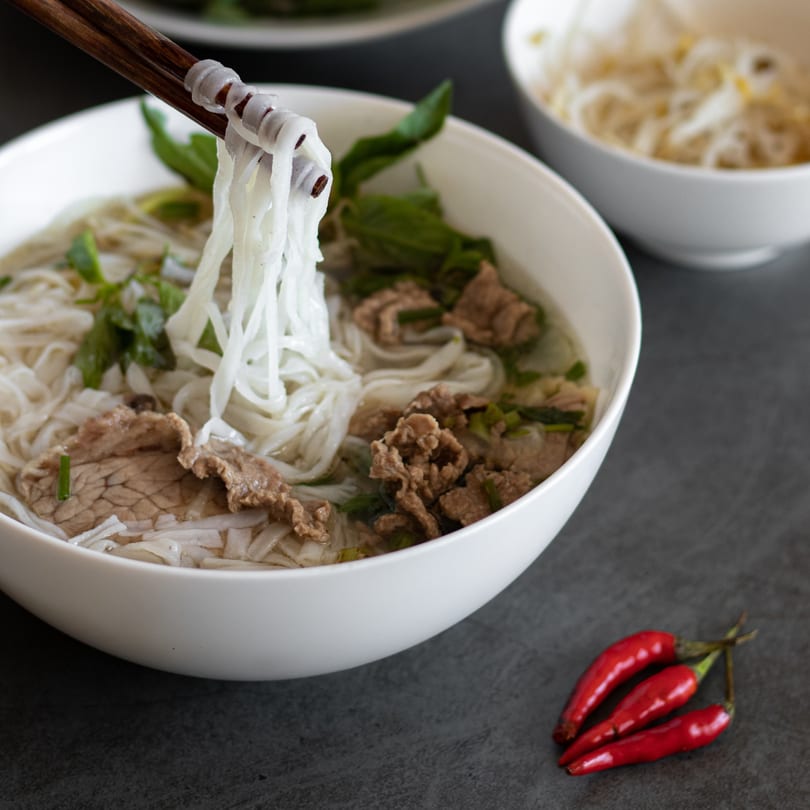 Chopsticks lifting noodles from a bowl of Chinese pho with beef, herbs, and red chilies.