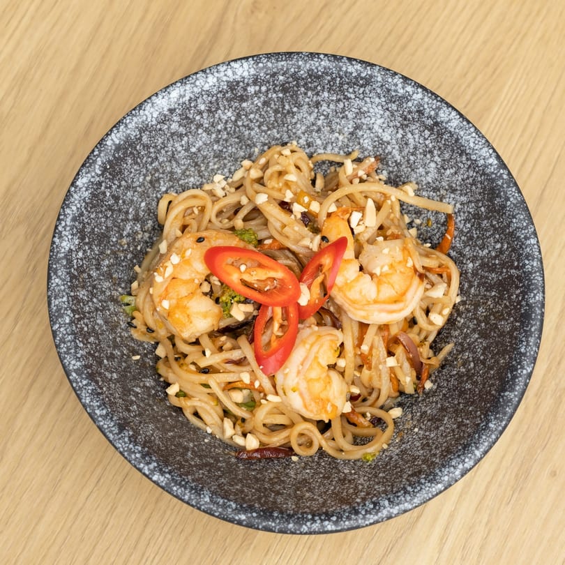 Lotus shrimp noodles with red chili slices, peanuts, and black sesame seeds in a dark bowl.